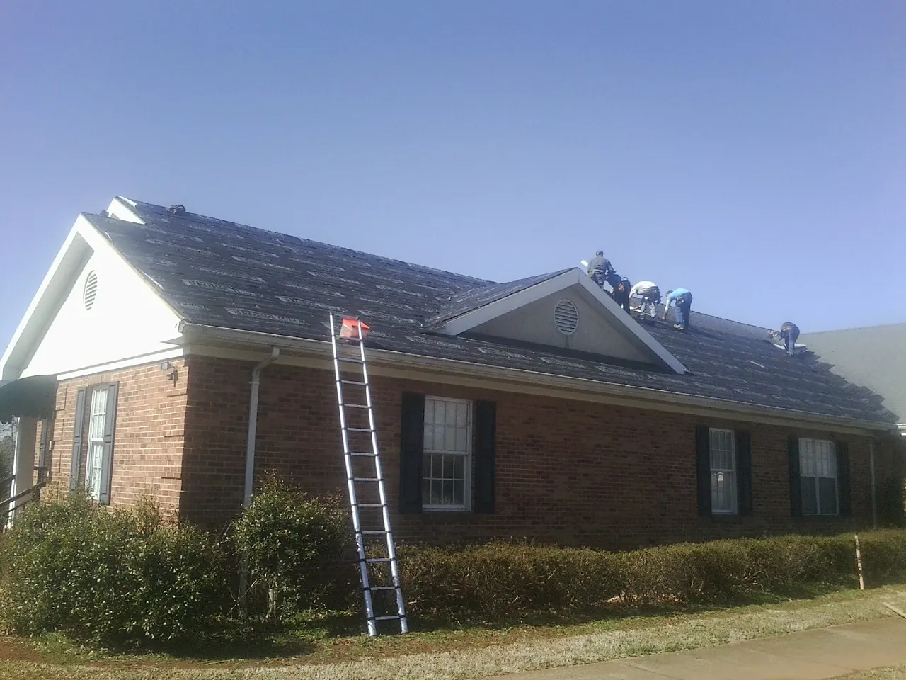 Crew tearing shingles off a brick ranch roof with a ladder set — Emergency Roof Repair work by our crew in Nevis